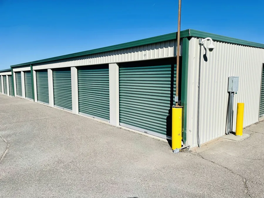 Outdoor self-storage building with a row of green roll-up doors, cream metal walls, yellow bollard and security camera on the corner, deep b